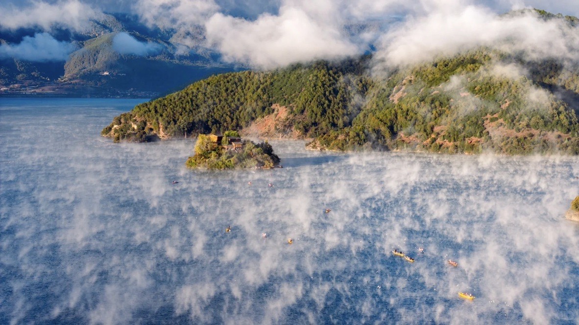 Forested island on Lugu Lake ringed by kayakers as mist rises off the water in golden morning light.