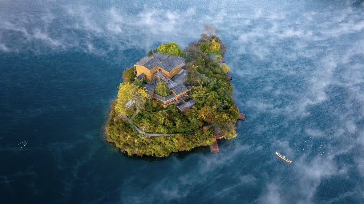 Aerial view of a small forested island crowned with wooden buildings, ringed by misty deep-blue water at Lugu Lake.