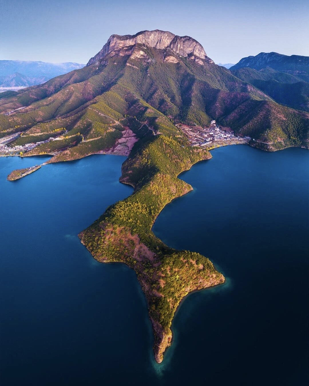 Aerial view of a wooded peninsula reaching into the deep blue water of Lugu Lake below a mountain.