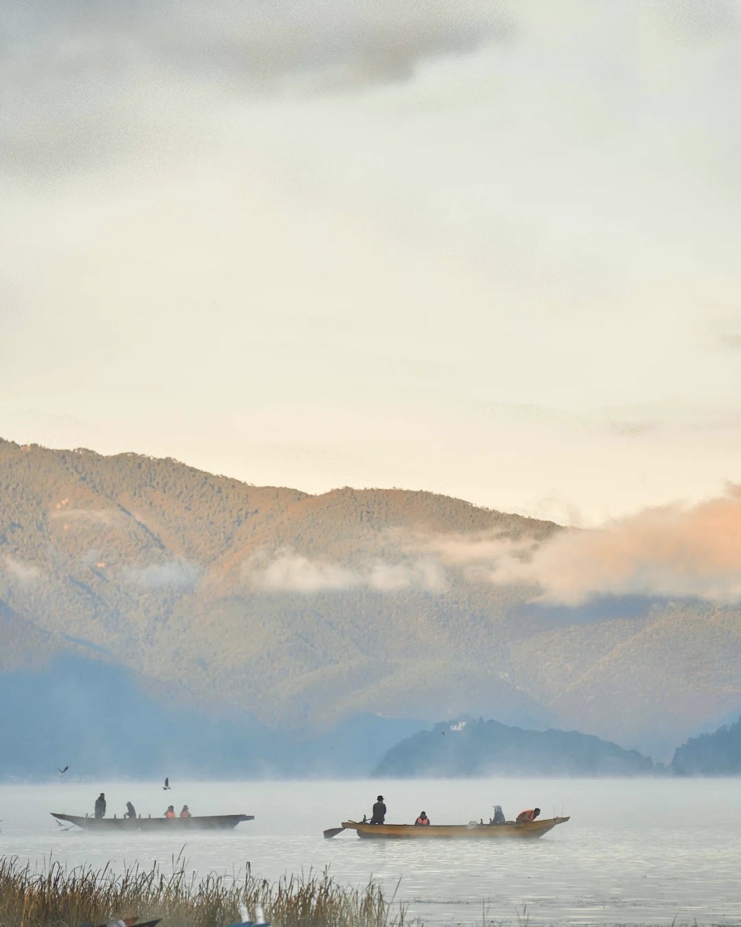 Two traditional wooden boats carry passengers across misty Lugu Lake at dawn below forested hills.