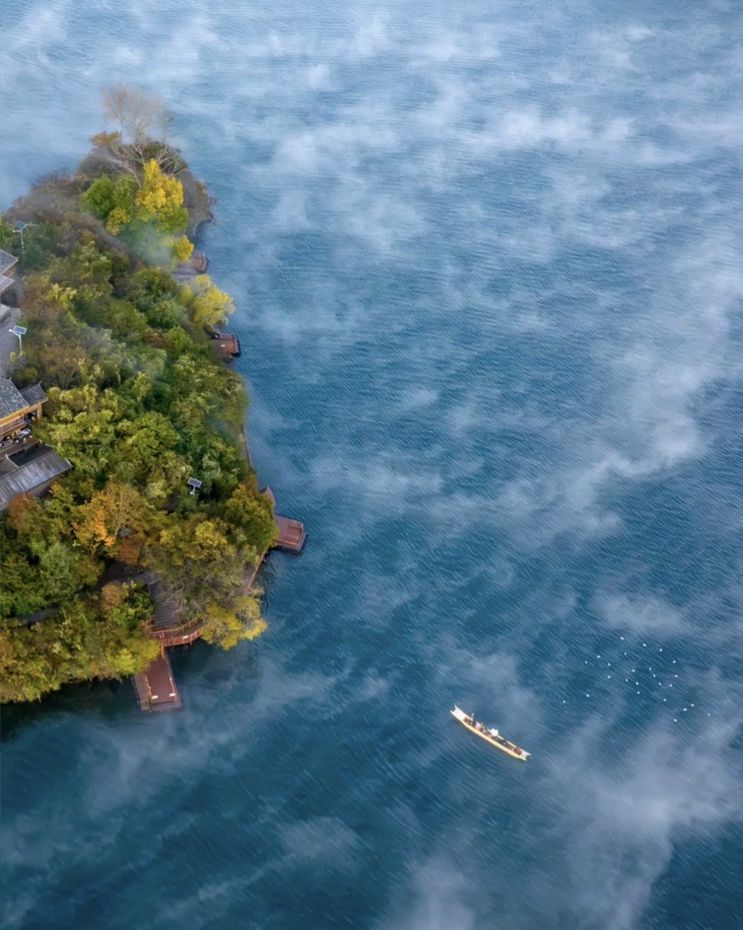 Aerial view of a wooded peninsula on Lugu Lake with mist drifting over blue water and a lone boat below.