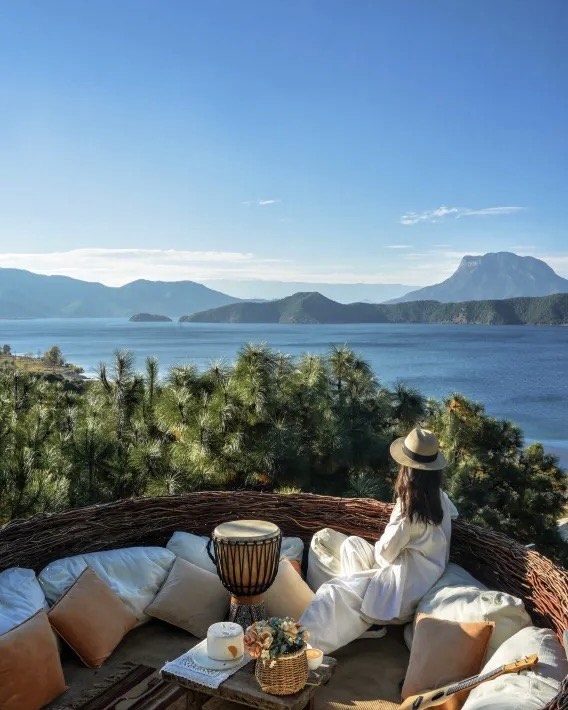 A woman in white sits on a cushioned terrace overlooking Lugu Lake's islands and mountains through pine trees.