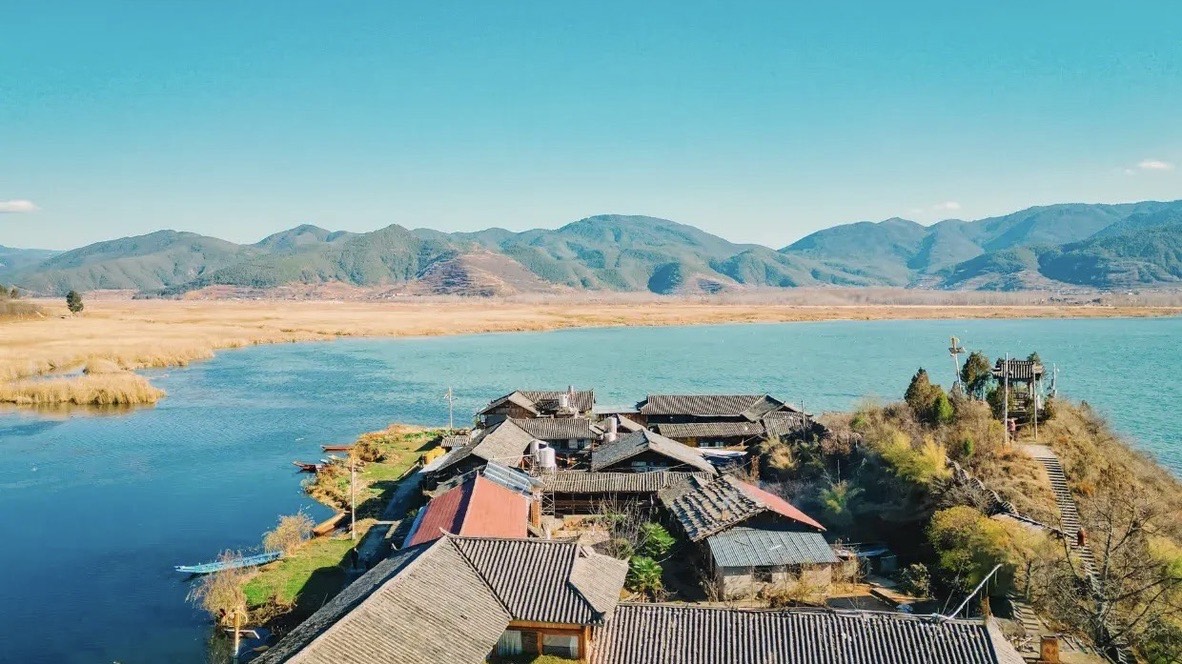 Aerial view of a lakeside village with grey-tiled roofs beside golden reeds and distant mountains at Lugu Lake.