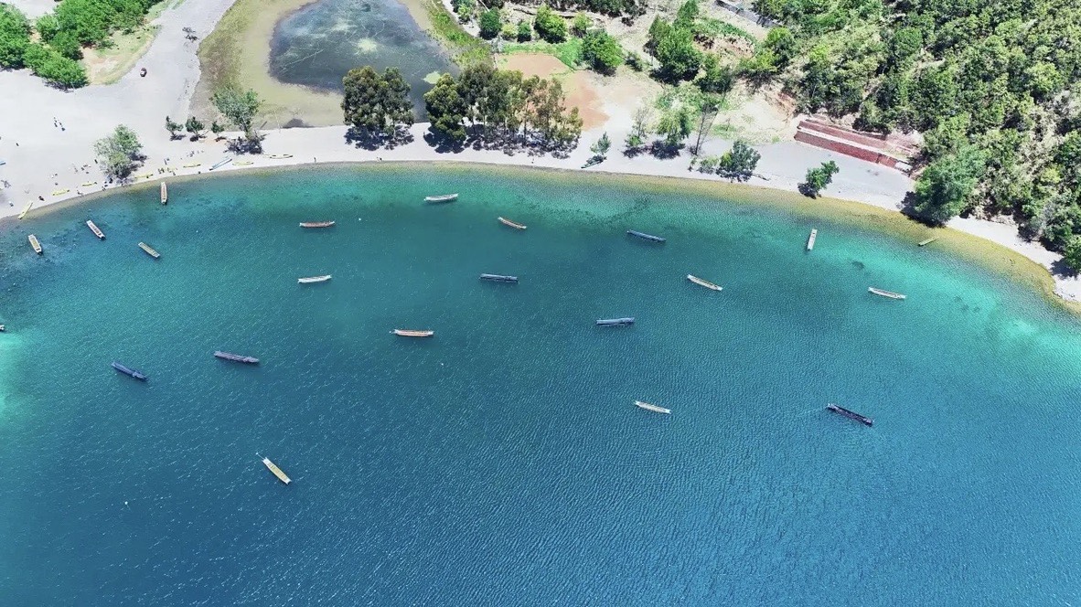 Aerial view of wooden boats moored across a clear turquoise cove fringed by a pine-lined shore at Lugu Lake.