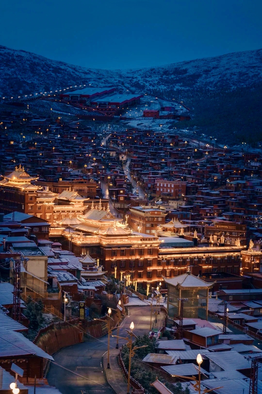 Seda's dense red Tibetan monastic houses climbing a snowy hillside, illuminated golden at dusk under a deep blue sky.