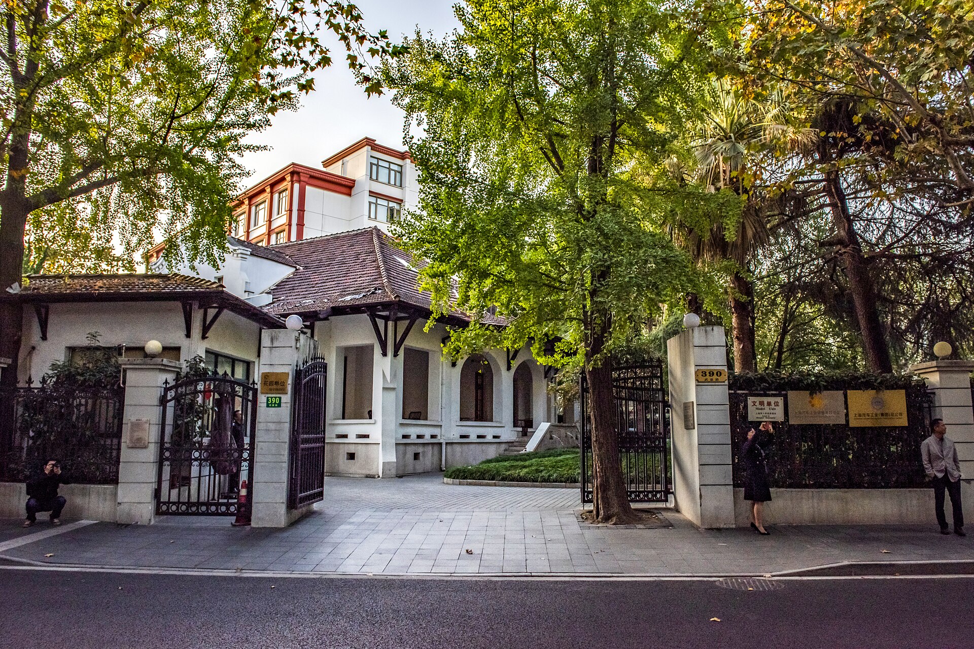 A historic villa gate on Wukang Road framed by lush green plane and ginkgo trees in warm afternoon light, former French Concession, Shanghai