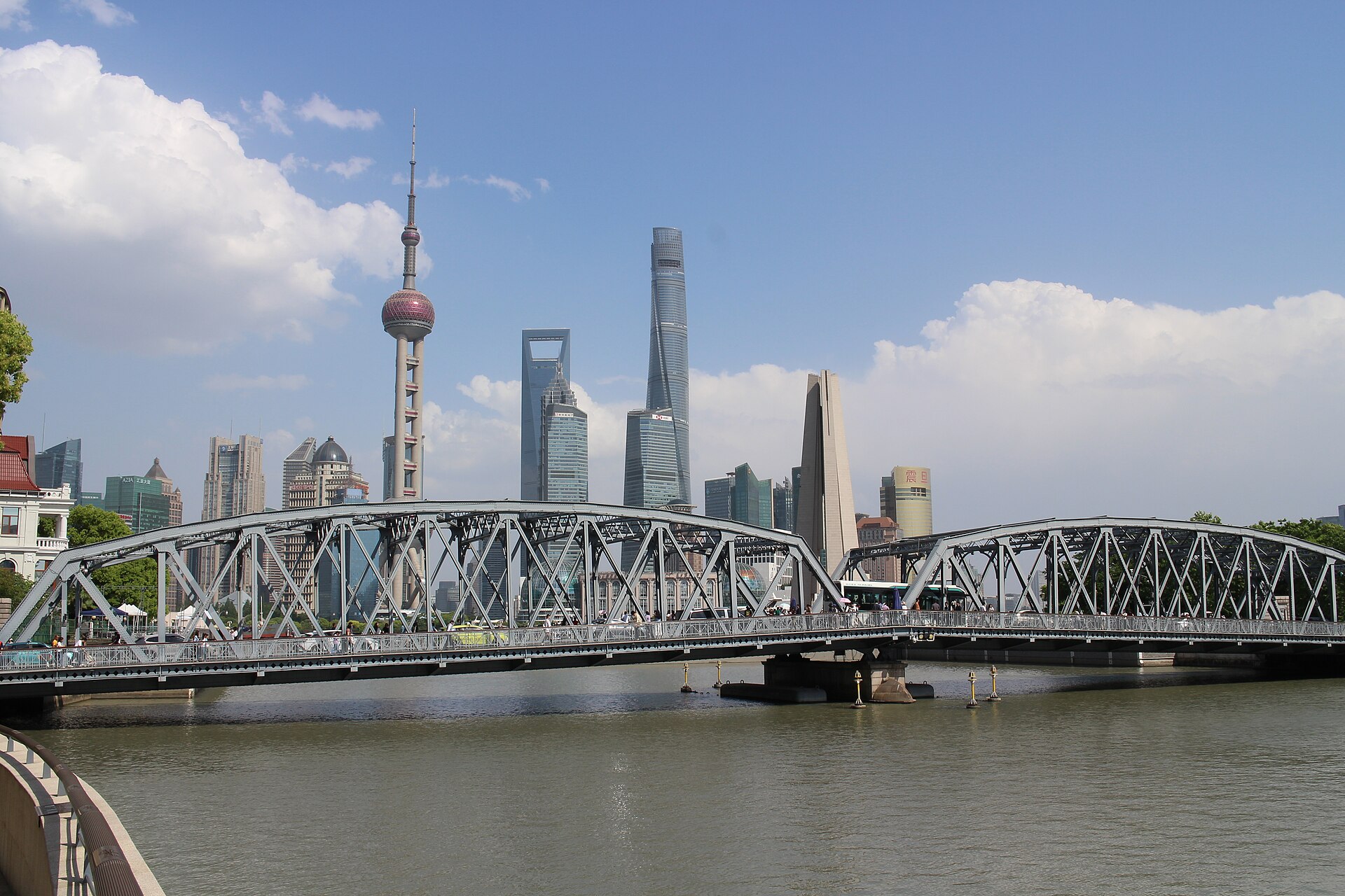 The silver steel trusses of Waibaidu Bridge (外白渡桥) arcing across Suzhou Creek under a blue summer sky, the Pudong towers beyond, Shanghai
