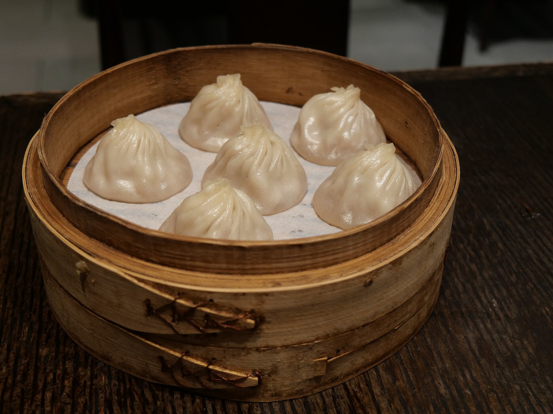 A bamboo steamer of soup dumplings (小笼包) on dark wood, their pleated tops glistening, the classic Shanghai plate