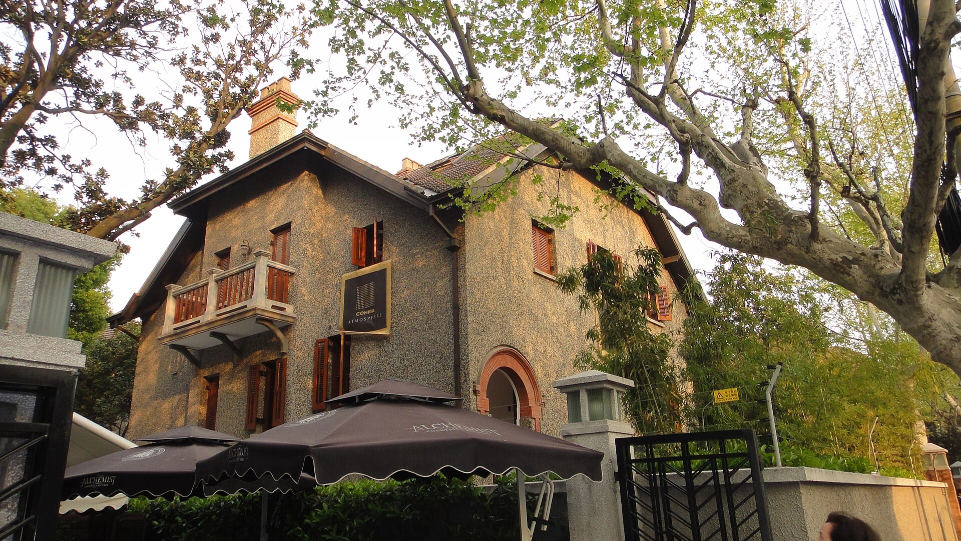 A stone-faced heritage villa with a café terrace under a leafy green plane tree on a quiet lane of the former French Concession, Shanghai