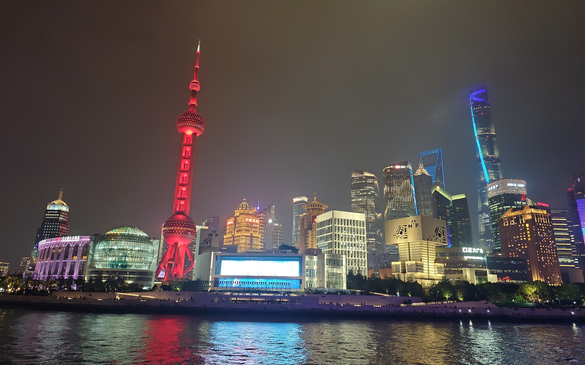 The Pudong skyline at night across the Huangpu River — the Oriental Pearl Tower (东方明珠) glowing beside the Shanghai Tower and Jin Mao cluster, seen from the Bund, Shanghai