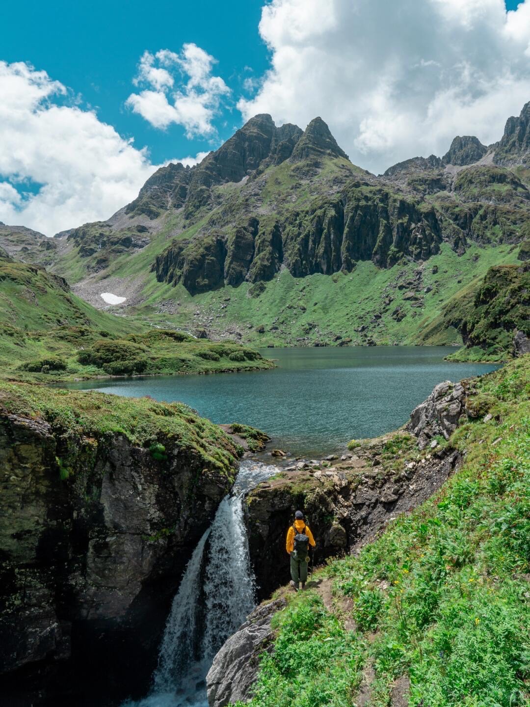 Hiker beside a waterfall and turquoise alpine lake below jagged green peaks on an off-beaten Shangri-La trek.