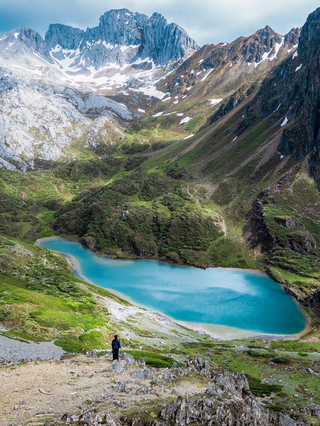 Solo trekker overlooks a turquoise alpine lake ringed by green slopes and jagged snow-dusted peaks on an off-beaten Shangri-la trek.