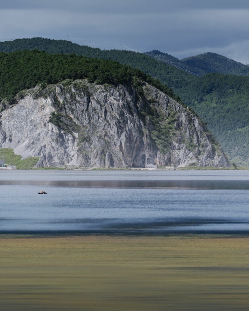 Rocky forested cliff rising above Napa Lake near Shangri-La, with a small boat on calm water and golden grassland in the foreground.