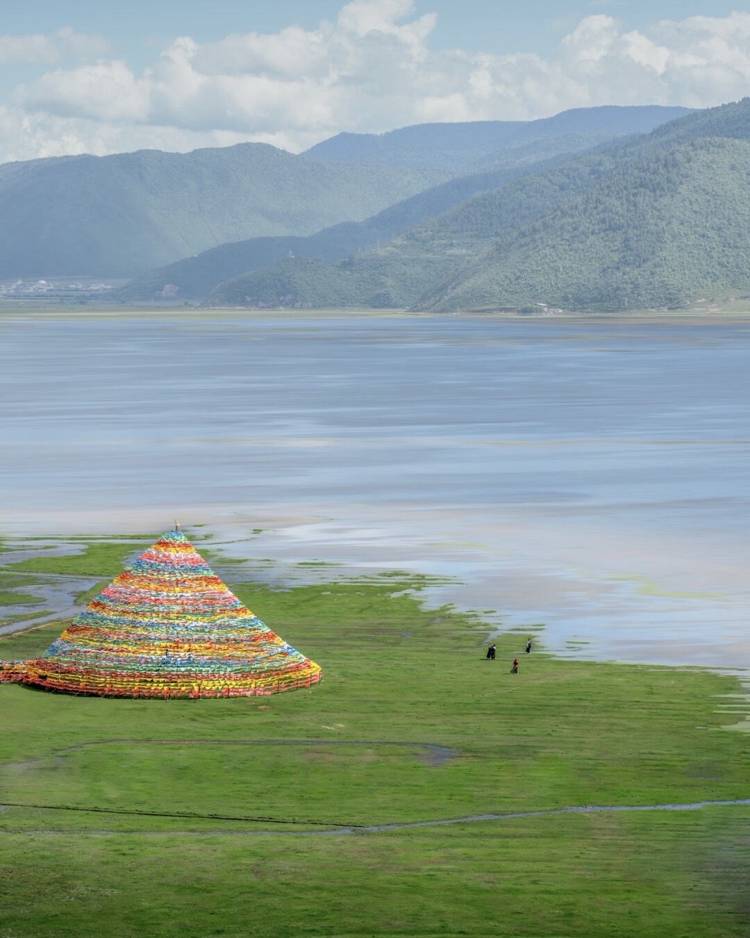 Colourful prayer-flag mound on the green grassland shore of Napa Lake near Shangri-La, with forested mountains beyond.