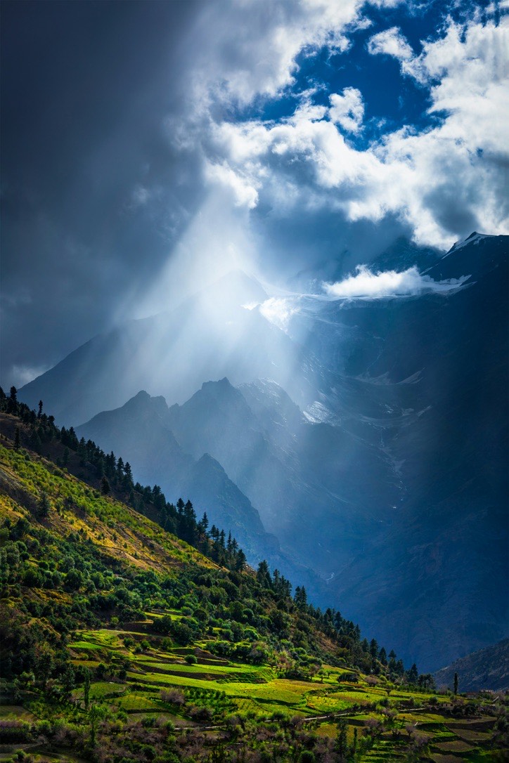 Sunbeams breaking through storm clouds over snow-dusted peaks above a terraced green valley near Shangri-La.