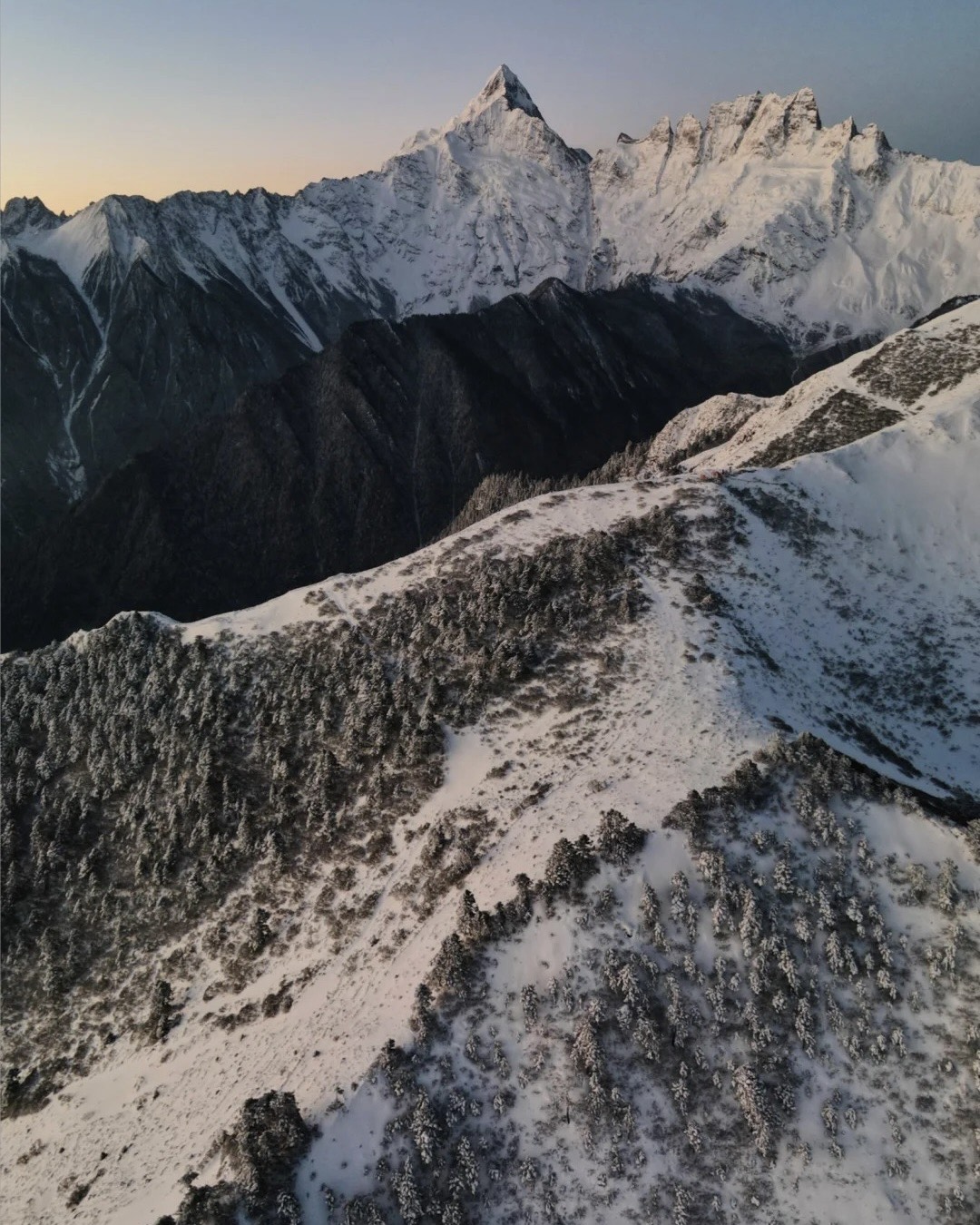 Aerial view of a snow-dusted forested ridge leading toward a sharp snow-capped peak at dawn on the Yubeng trek near Shangri-La.