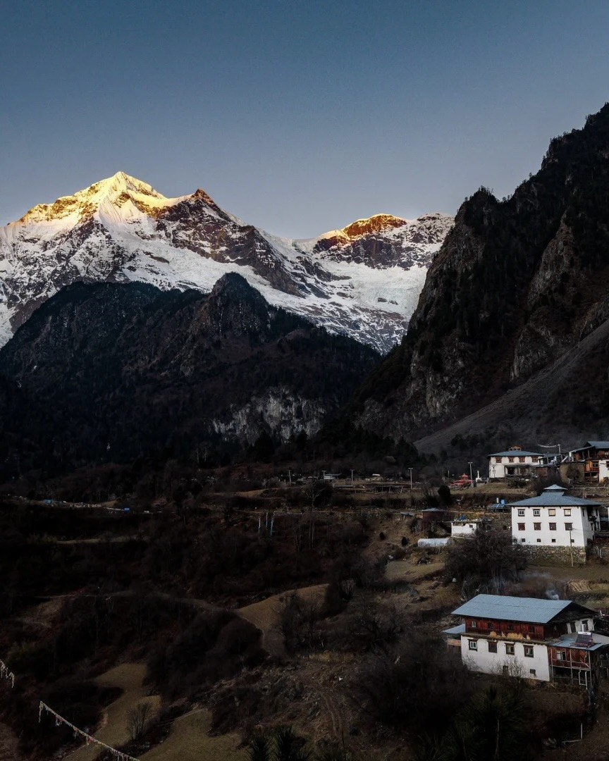 First light striking snow peaks above Yubeng village, with Tibetan whitewashed houses clustered in the valley at dusk near Shangri-La.