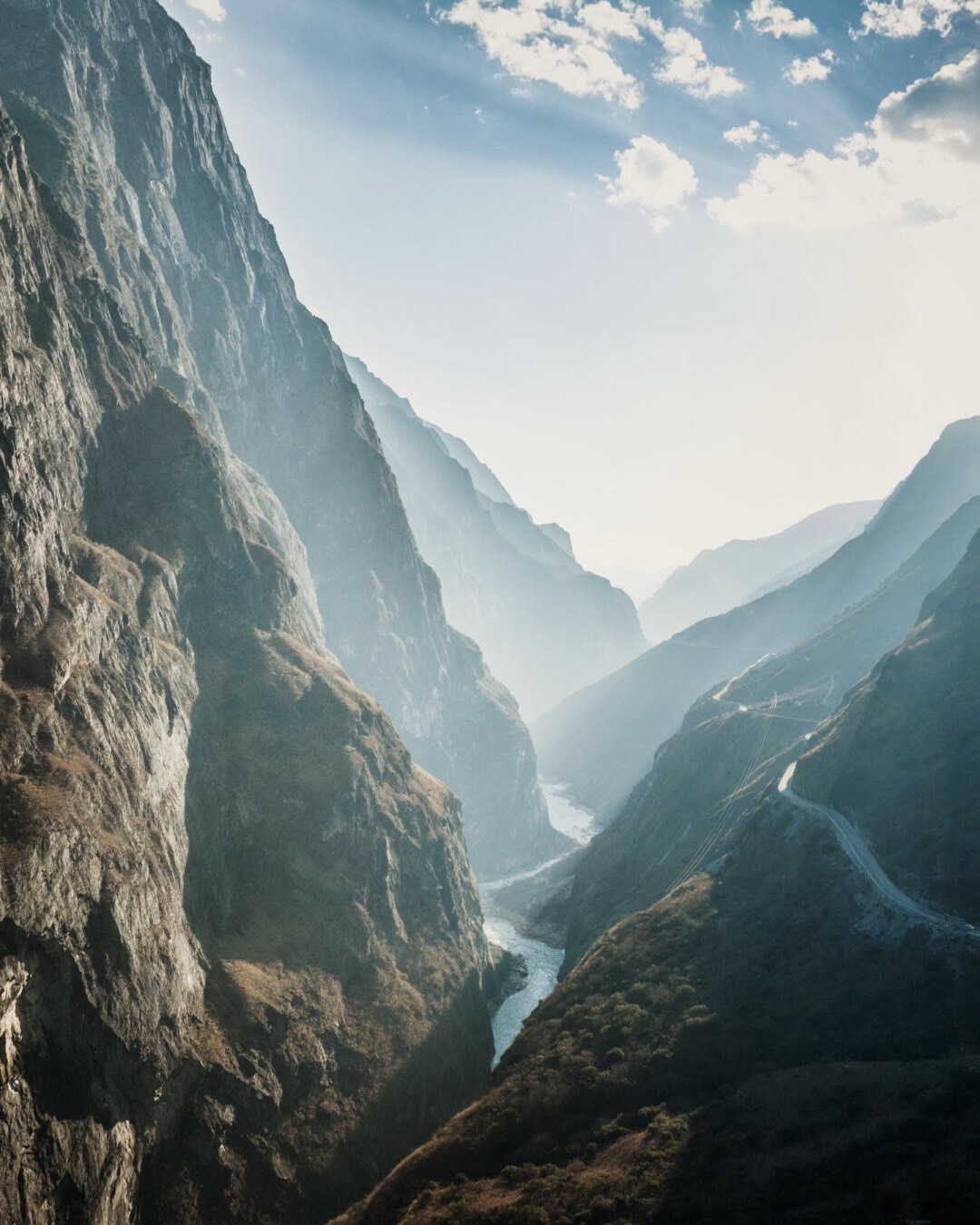 Deep river canyon of Tiger Leaping Gorge winding between towering cliffs into hazy sunlit ridges, with a road cut along the far slope.