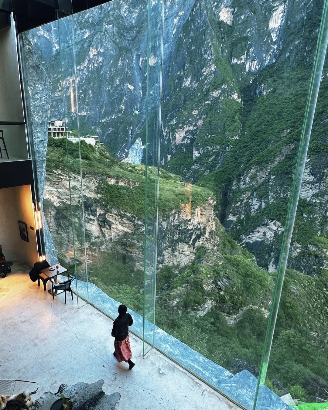 Guest walks past a towering glass wall framing the green cliffs of Tiger Leaping Gorge from a lounge overlooking the canyon.
