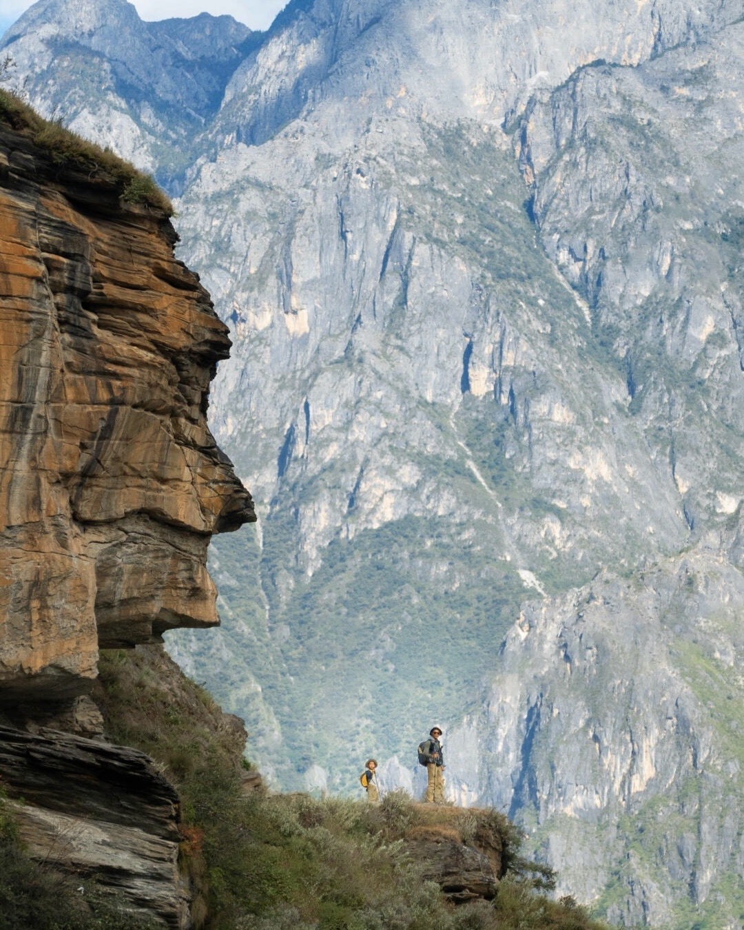 Two hikers standing on a narrow cliff ledge dwarfed by towering rock walls in Tiger Leaping Gorge near Shangri-La.