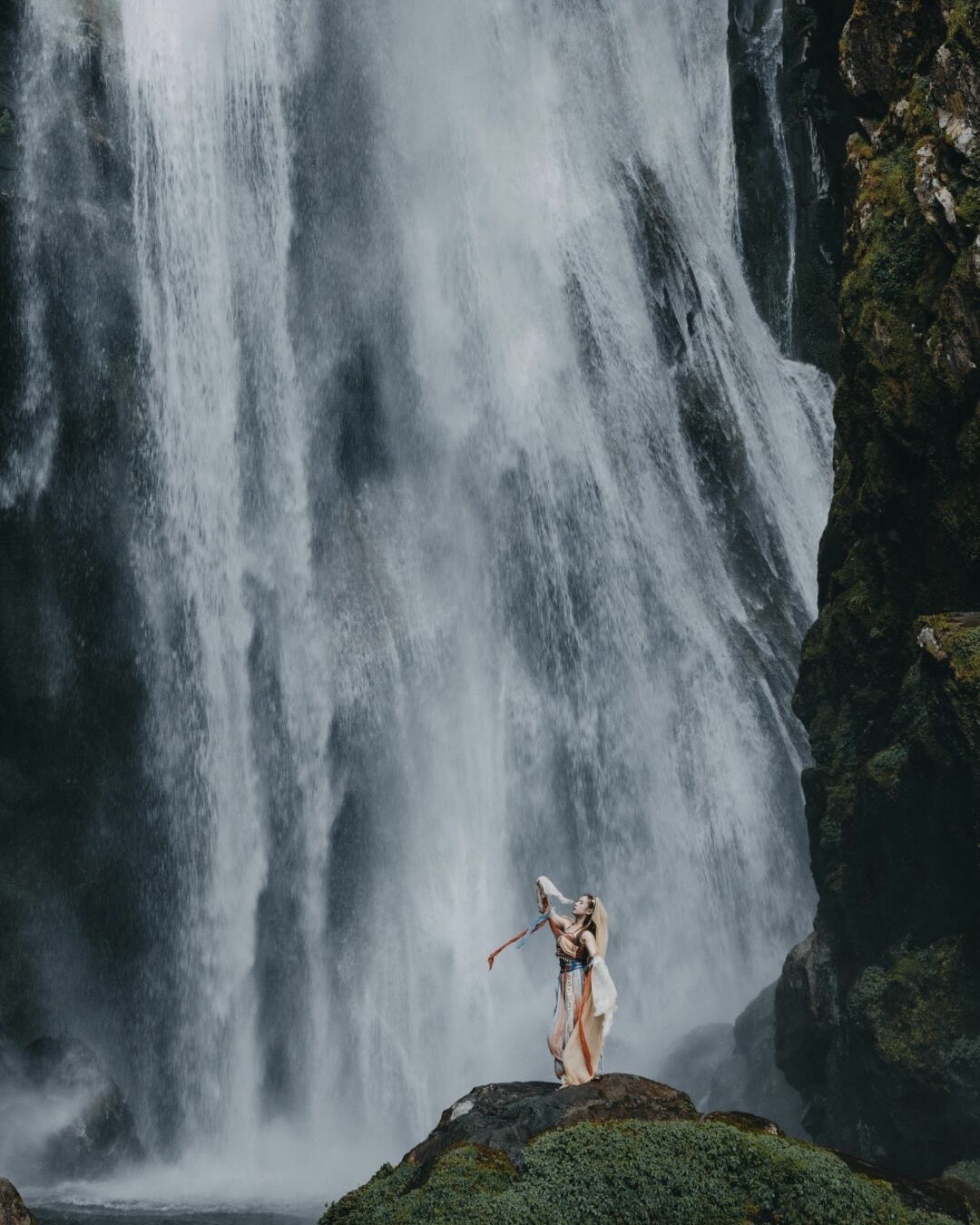 A dancer in flowing costume with trailing ribbons poses on a mossy rock before a tall waterfall near Gaoligong.