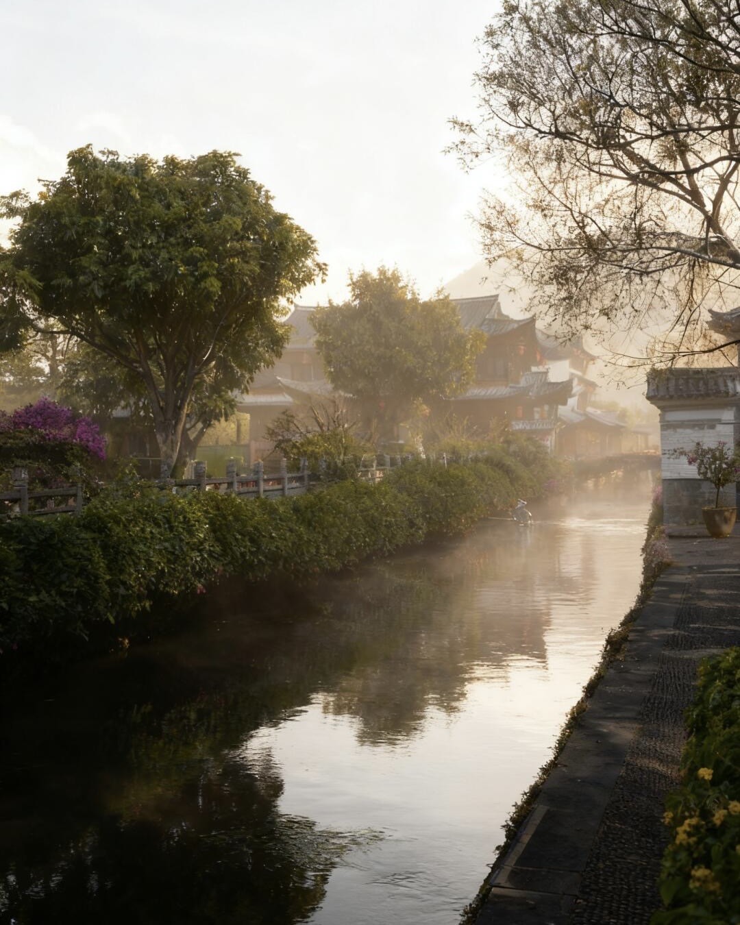 Golden misty sunrise over a canal lined with hedges and tiled-roof houses in Heshun ancient town.