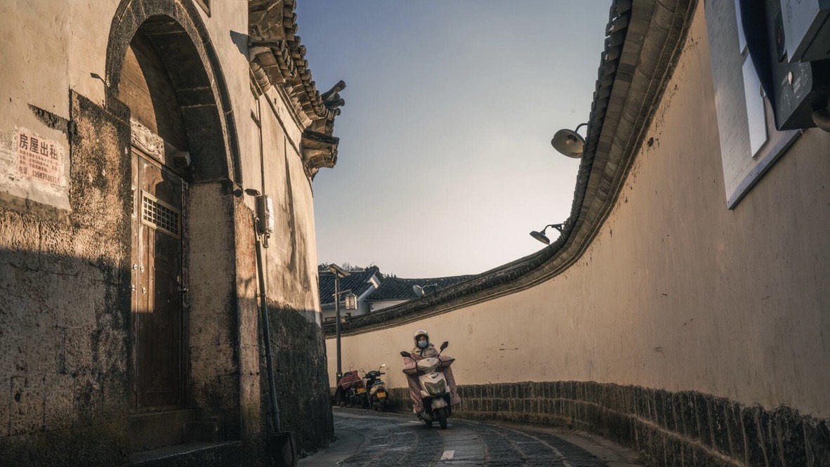 A scooter rider climbs a curved stone lane between rammed-earth walls and an old archway in Heshun ancient town.