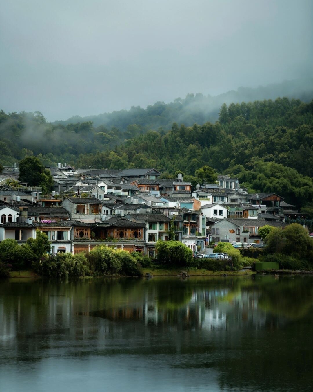 Tiered grey-tiled houses of Heshun ancient town reflected in a still lake below misty forested hills.