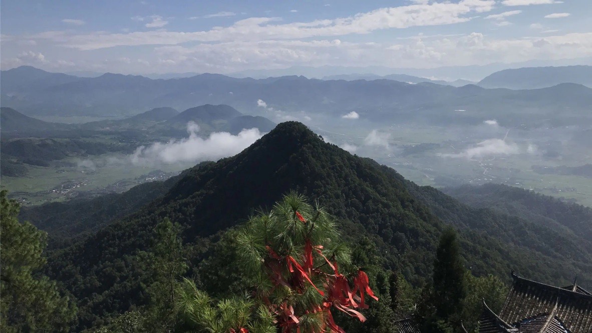 A conical forested peak rising above a hazy patchwork valley, with red prayer ribbons tied to a pine in the foreground.