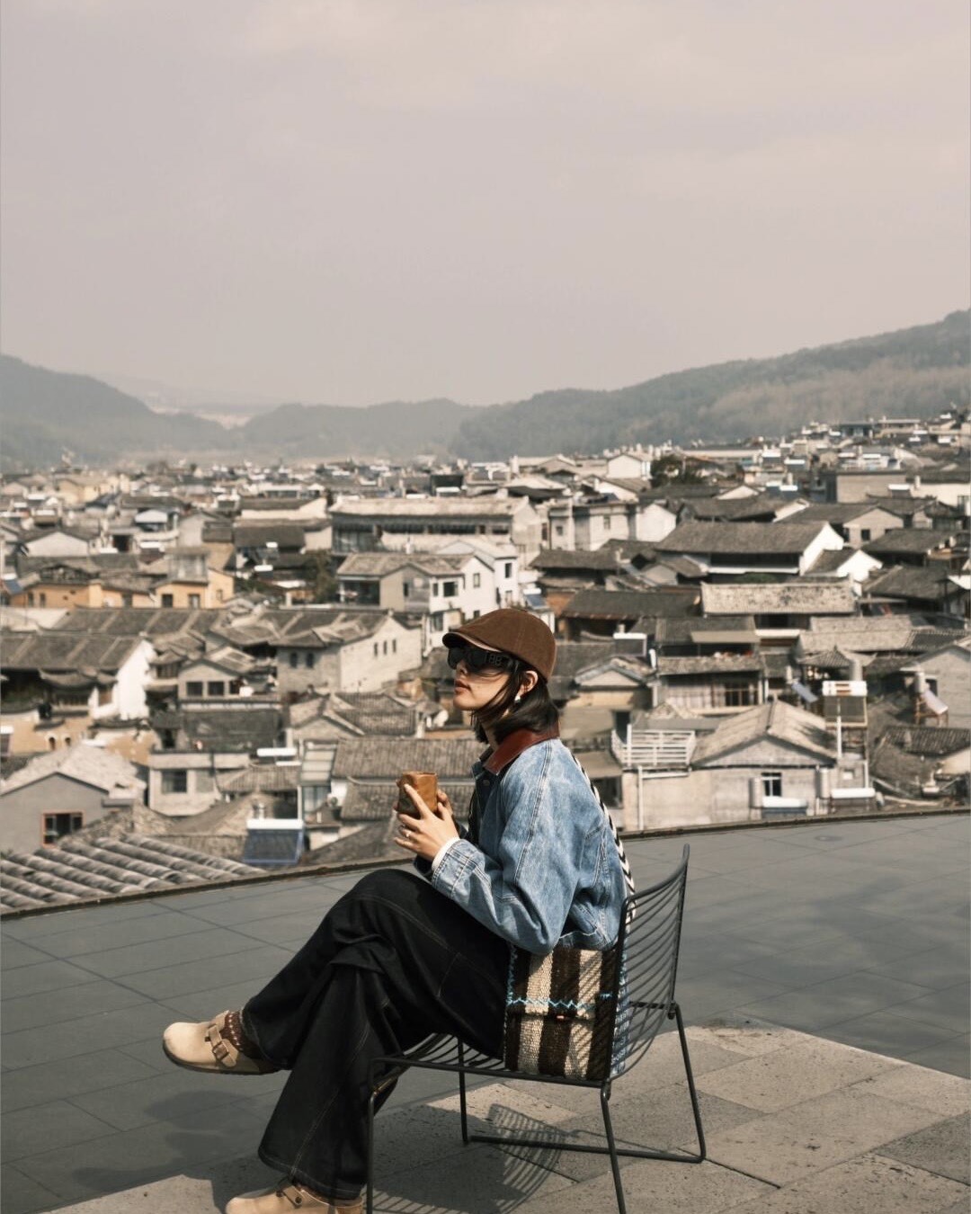 Traveller seated on a rooftop terrace overlooking the tiled roofs of Heshun Ancient Town and distant hills in Tengchong.