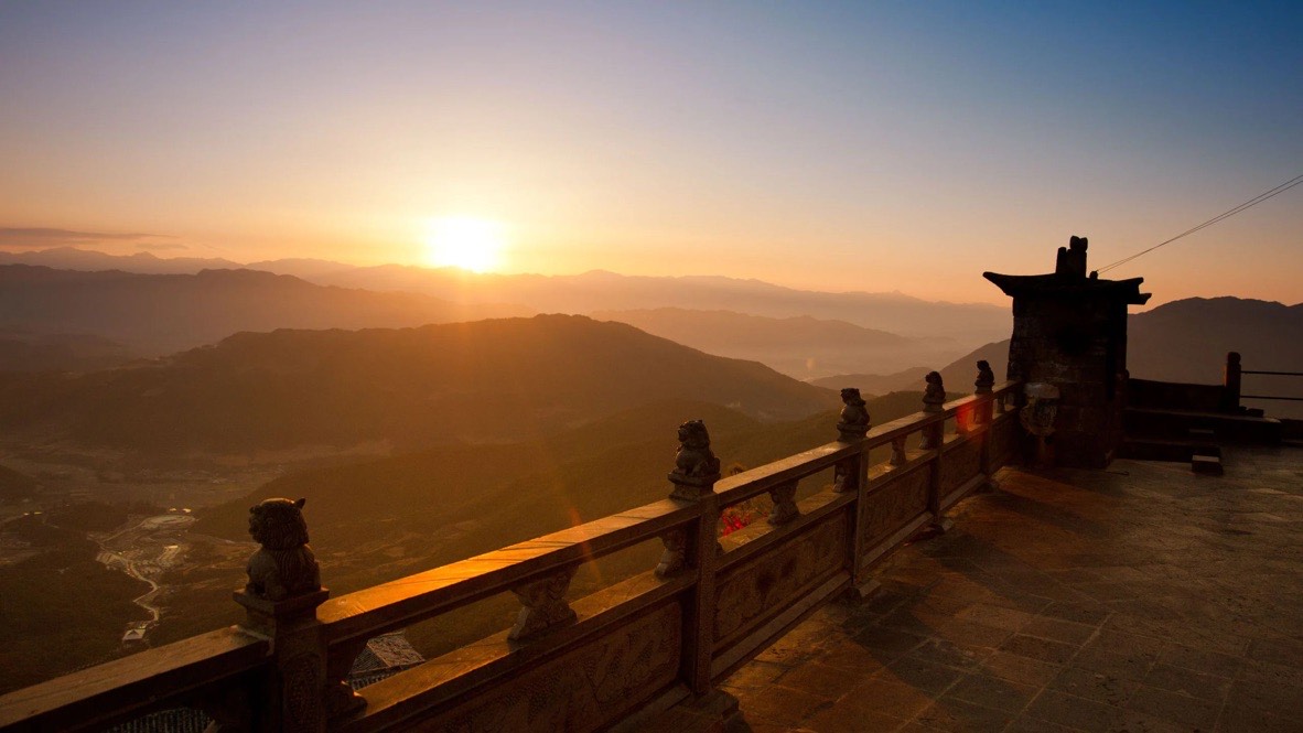 Sun rising over layered misty mountains beyond a stone temple terrace lined with carved lion balustrade figures.