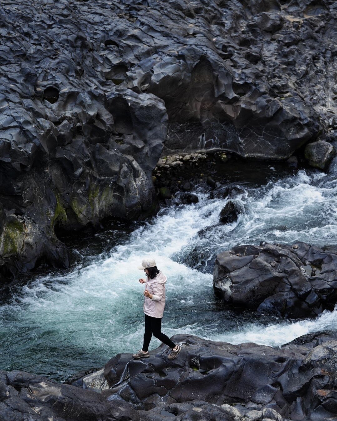 A hiker in a cap steps across dark basalt boulders beside churning white rapids in a Tengchong river gorge.