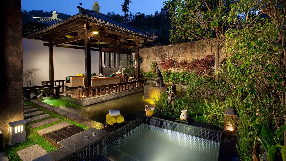 Lantern-lit hot-spring courtyard with a tiled soaking pool and timber pavilion at dusk near Tengchong's Rehai.