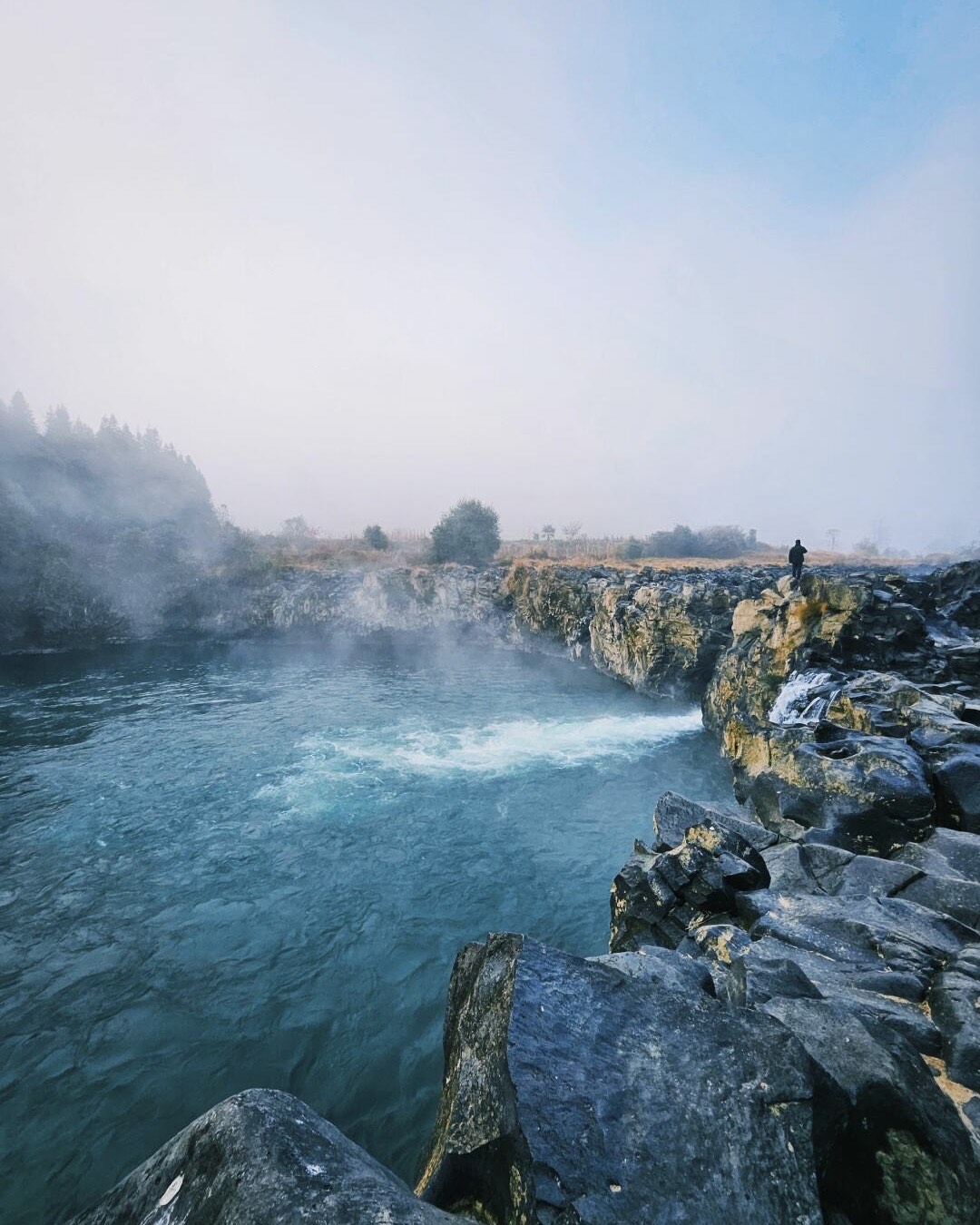 Steam drifts over a turquoise hot-spring pool ringed by dark basalt rock with a figure on the far ledge at Rehai.