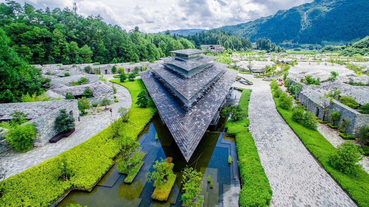 Aerial view of a stepped slate-roofed pyramid building set among reflecting pools and green gardens ringed by forested hills in Tengchong.