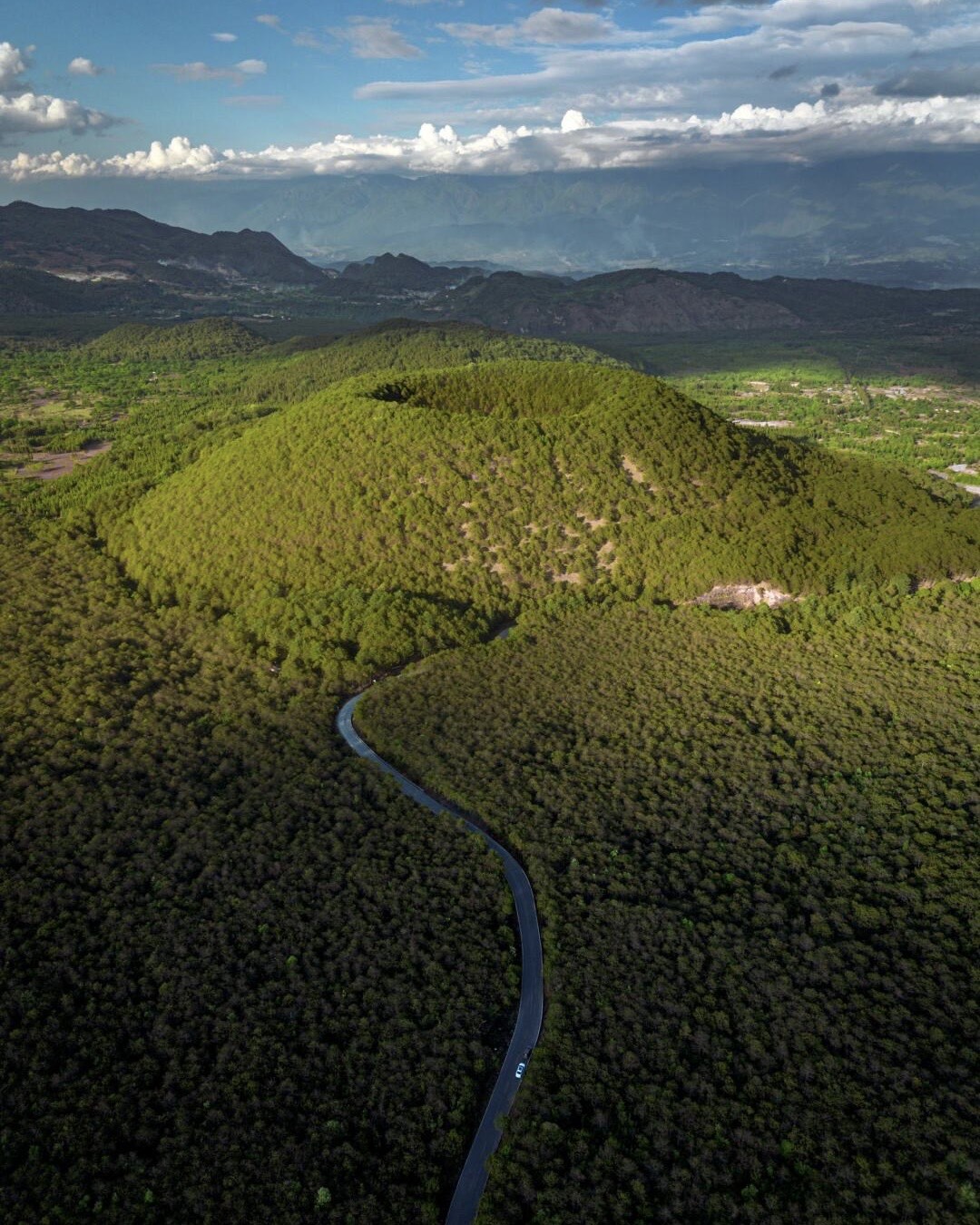 Aerial view of a forest-covered volcanic crater in Tengchong with a winding road threading through the green woodland.