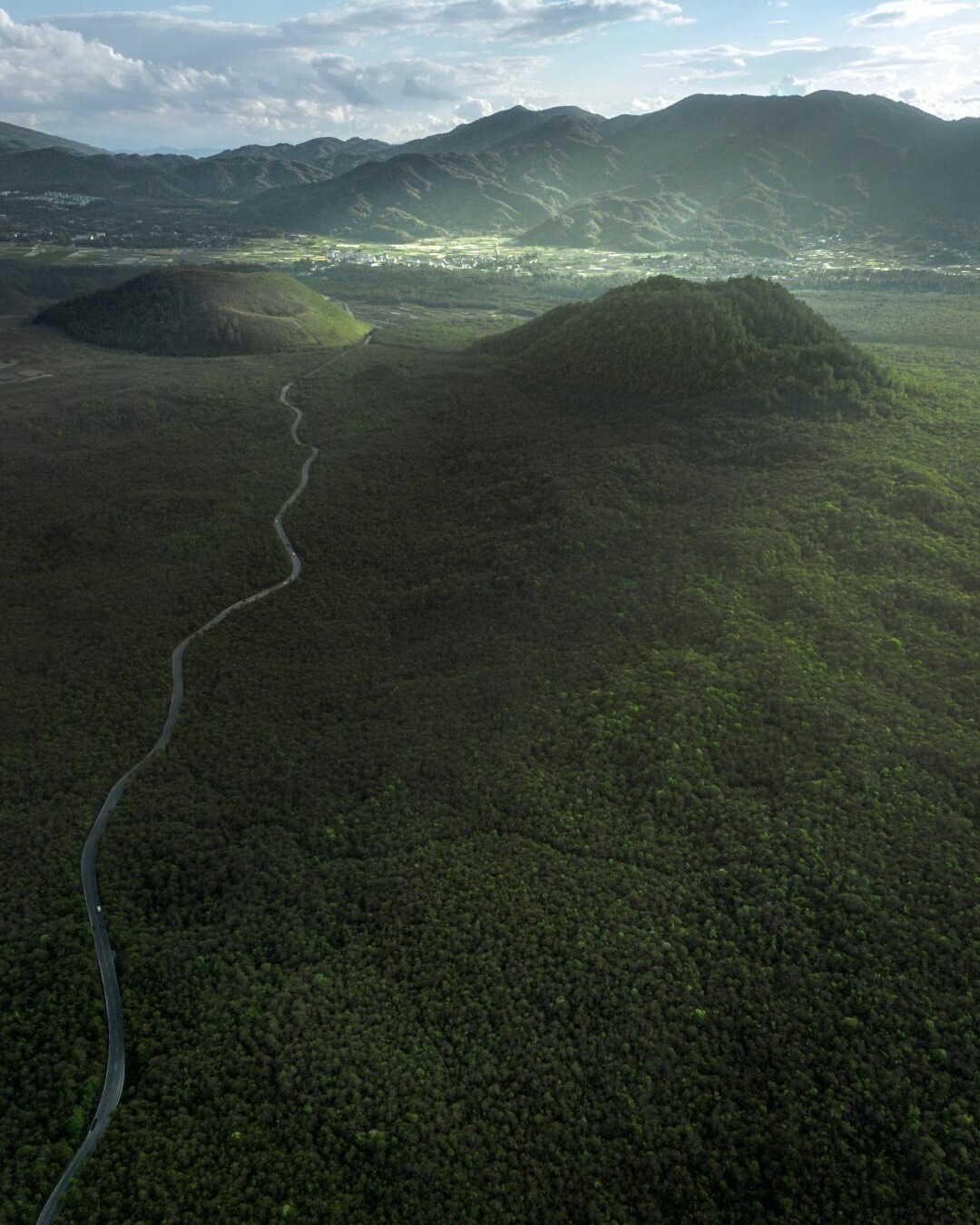 Aerial view of two forest-covered volcanic cones and a winding road across the plain at Tengchong's volcano park.