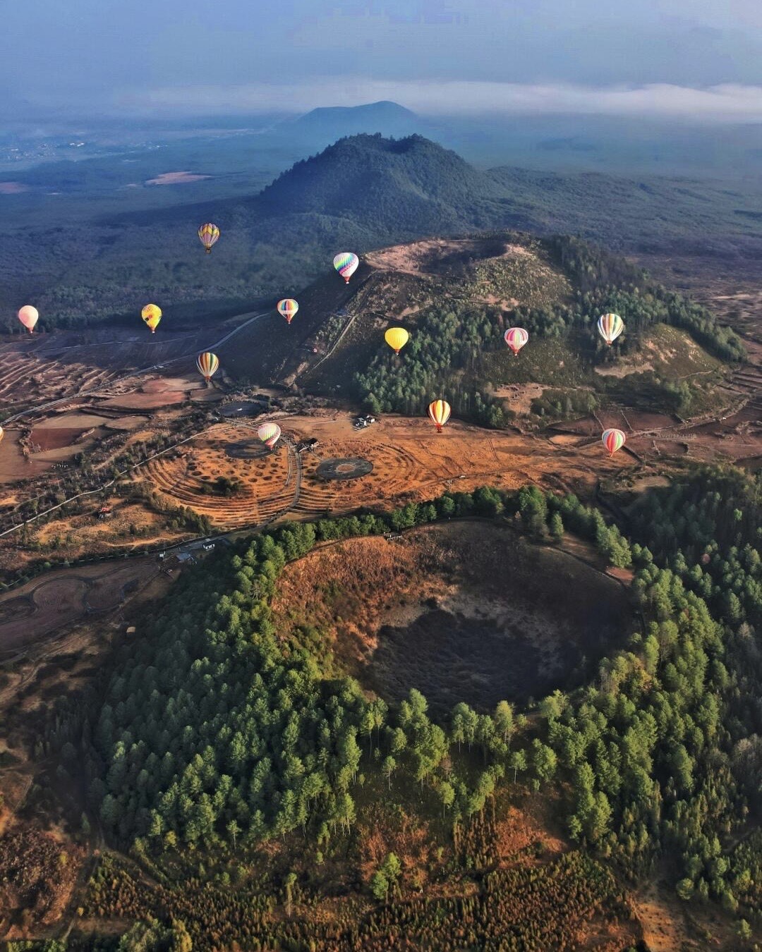 Aerial view of colourful hot-air balloons drifting over forested volcanic craters and cones in a wide valley.