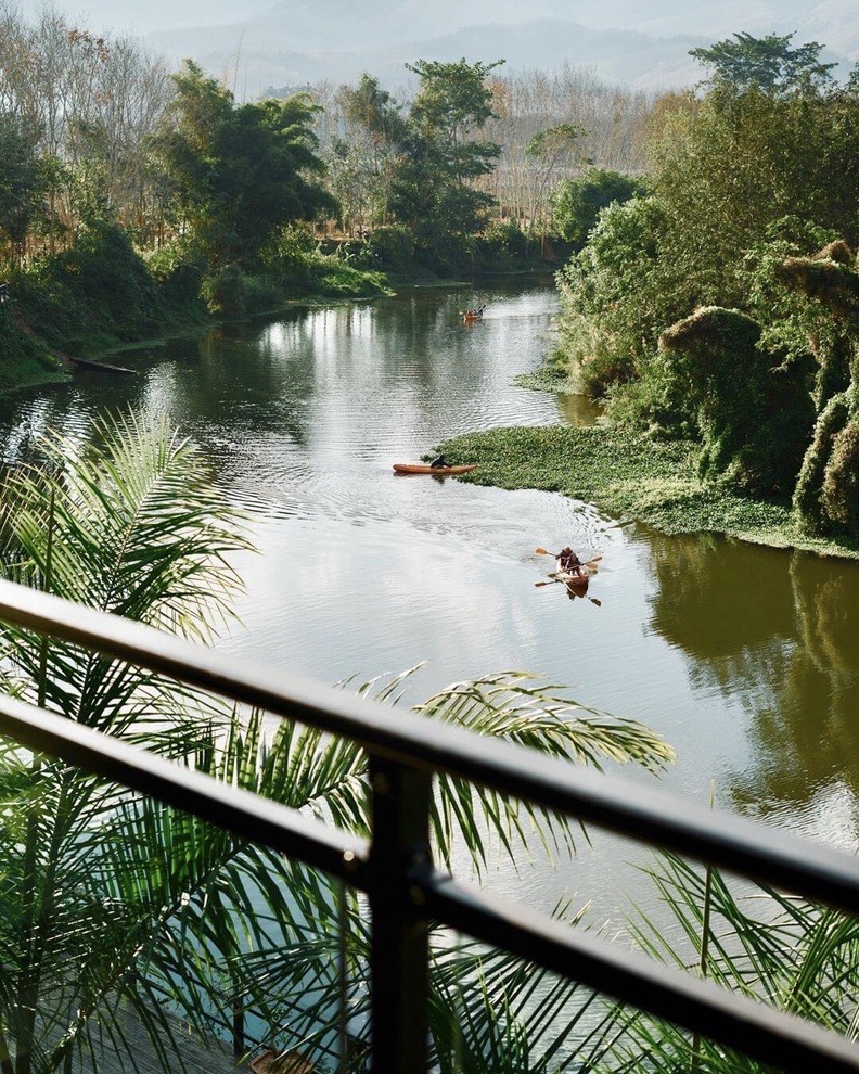 Kayakers paddle a calm river fringed by tropical greenery and distant hills, seen past a balcony railing.