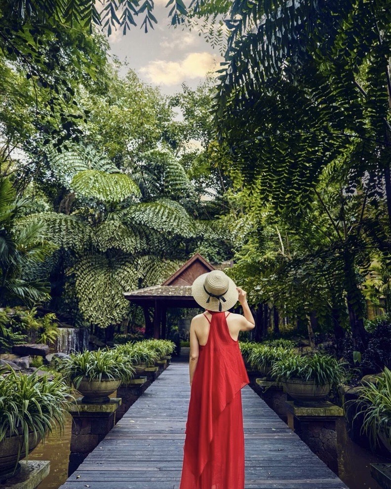 Woman in a red dress and straw hat on a wooden boardwalk lined with potted plants and tall tree ferns.