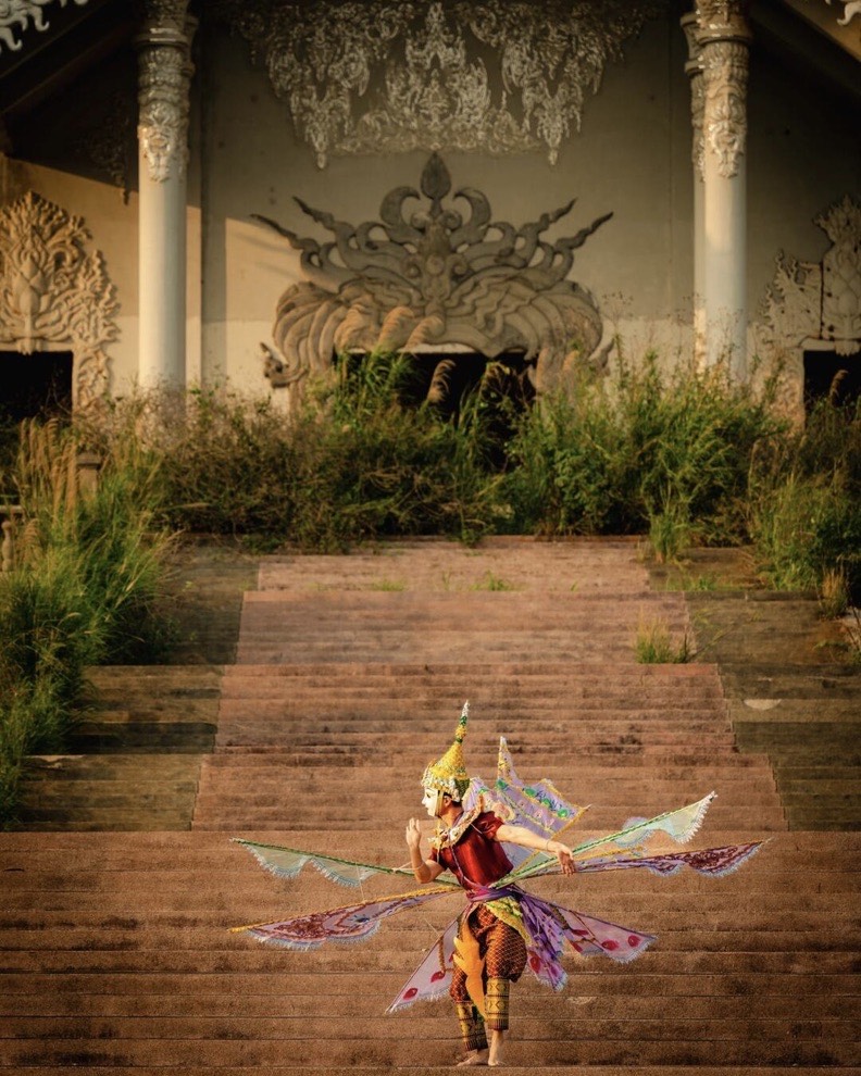 A costumed dancer in an elaborate winged headdress performs on wide temple steps at golden hour.