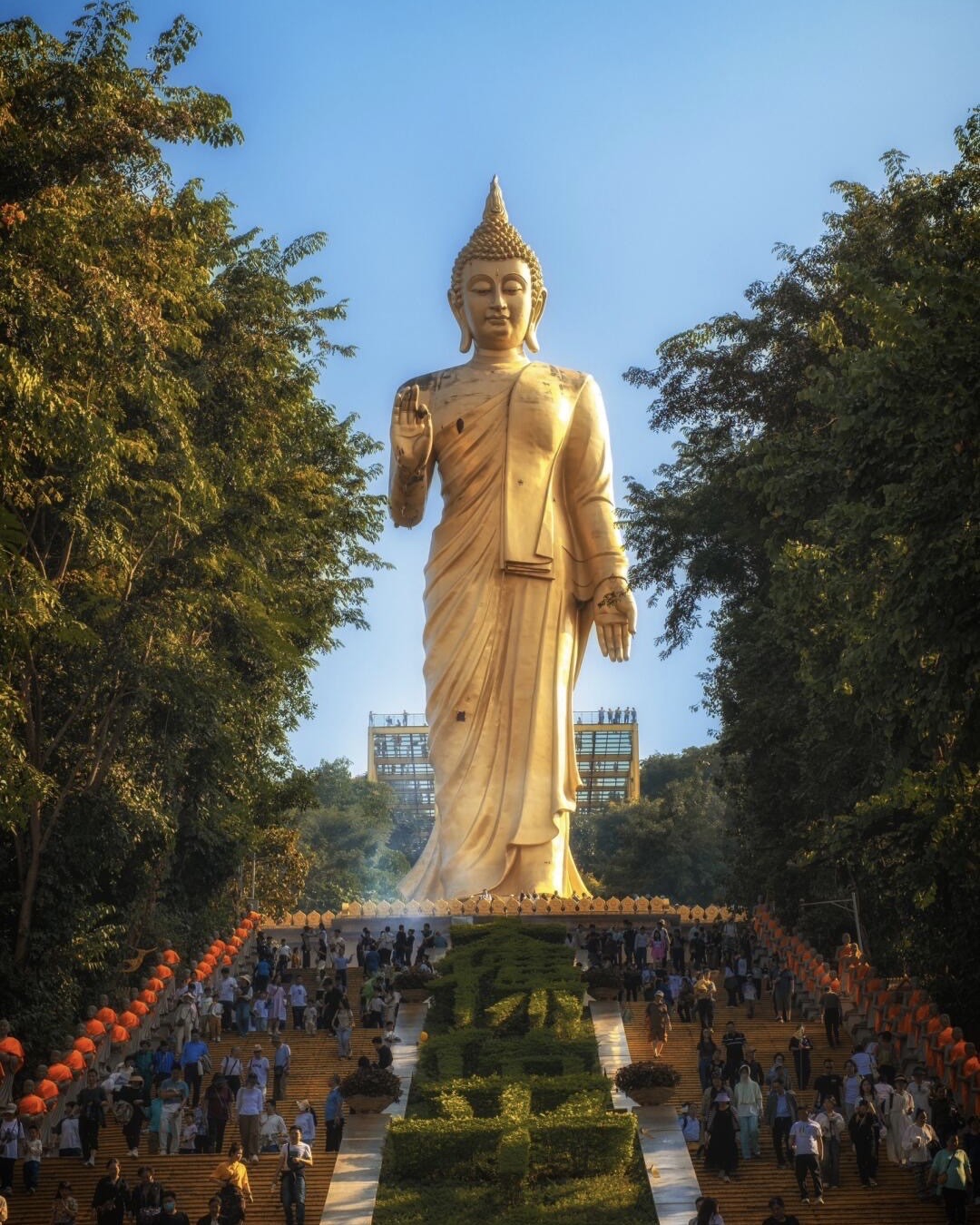 Towering golden standing Buddha statue above a long staircase lined with lanterns and crowds of visitors under blue sky.