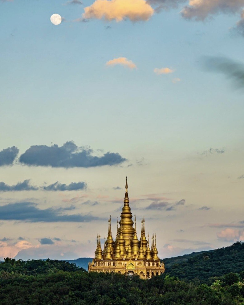 Golden multi-spired pagoda on a forested hilltop beneath a pastel evening sky with the moon rising above.
