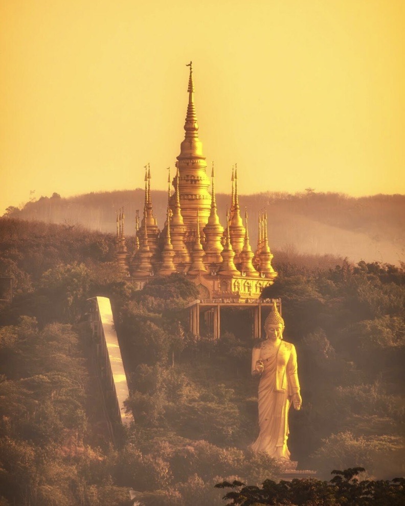 Golden spired pagoda cluster and a tall standing Buddha statue on a forested hillside glowing in warm sunset haze.