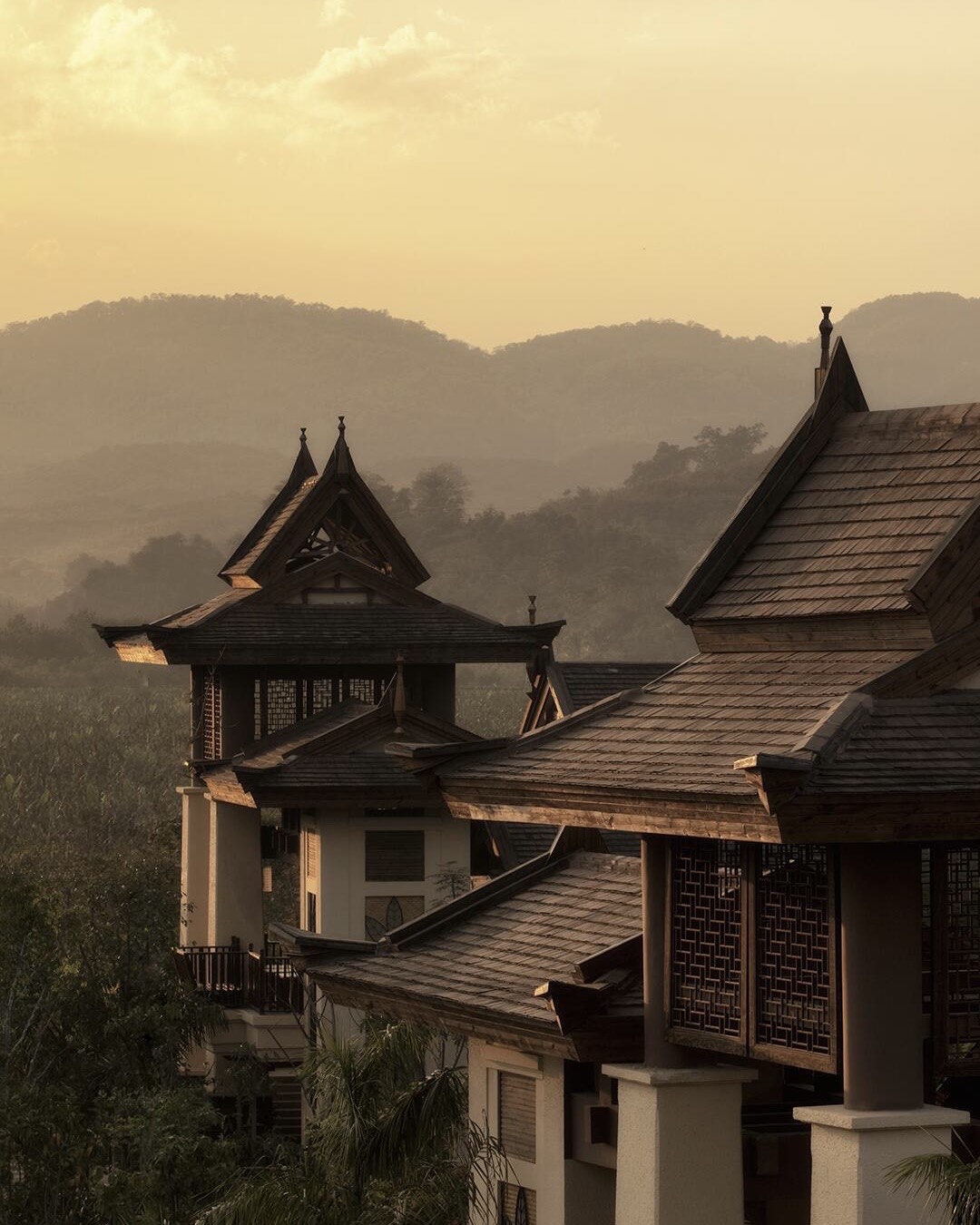 Layered wooden Dai-style pavilion roofs with carved lattice windows against misty forested hills at golden dusk.