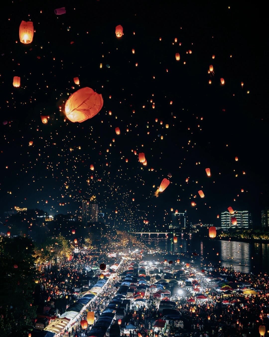 Hundreds of glowing sky lanterns rising over a crowded riverside night market lit up after dark.