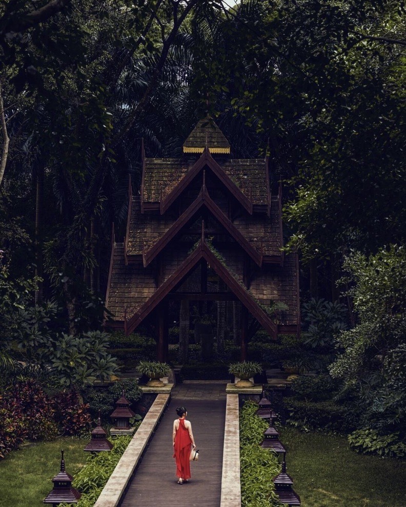 Woman in a red dress walks a garden path toward a steep tiered wooden temple set in dense jungle.