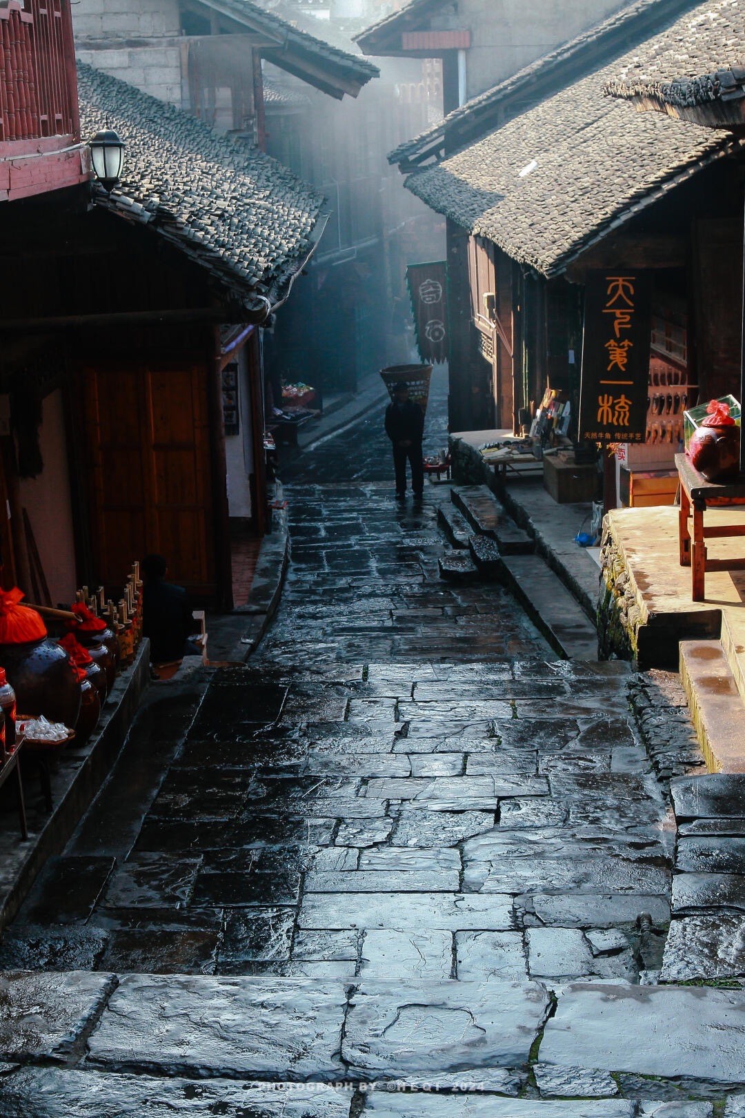 Rain-slicked flagstone lane between old wooden shopfronts with a porter carrying a back-basket in early light.