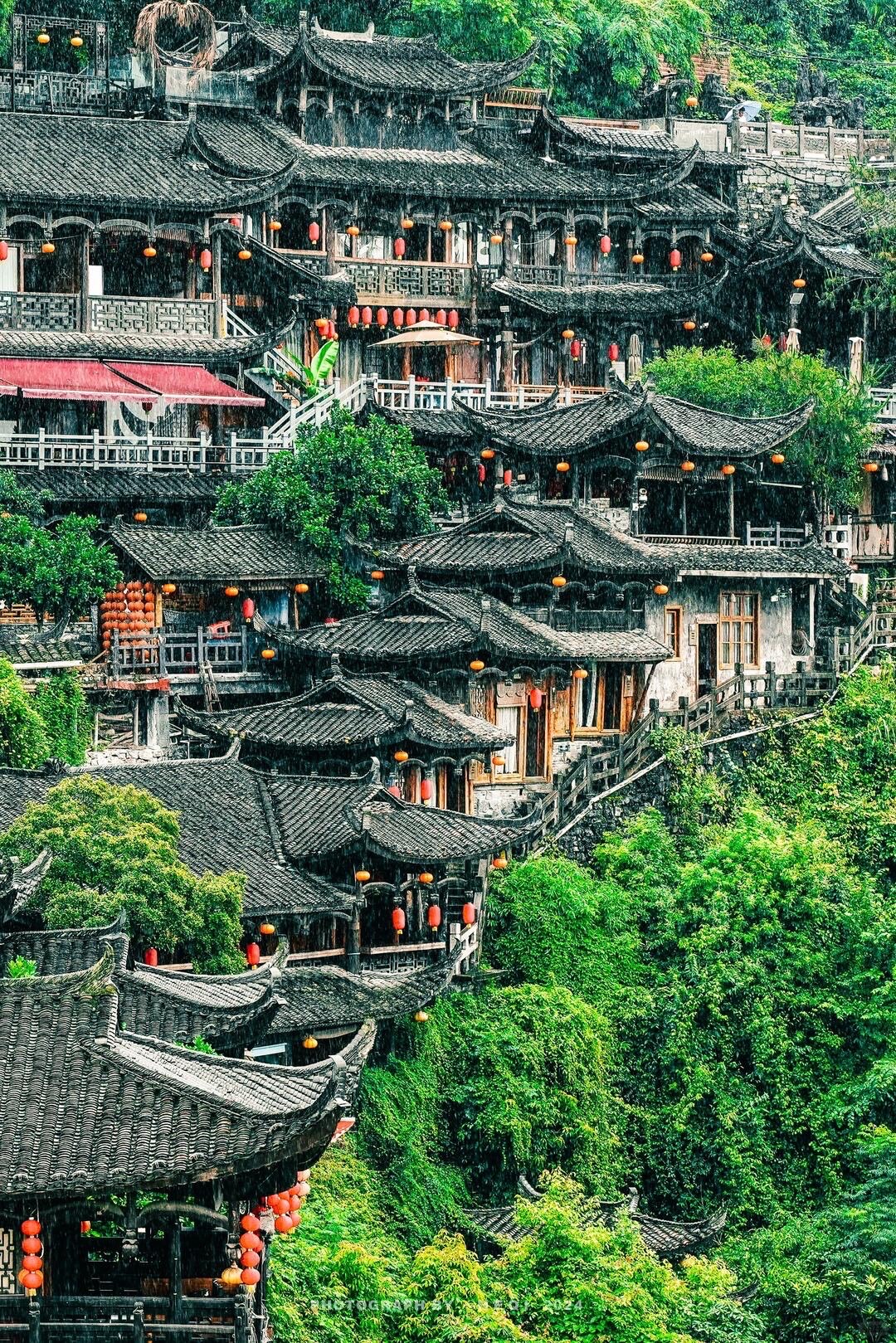 Densely stacked tiered black-tiled rooftops hung with red lanterns clinging to a green cliffside in the rain.