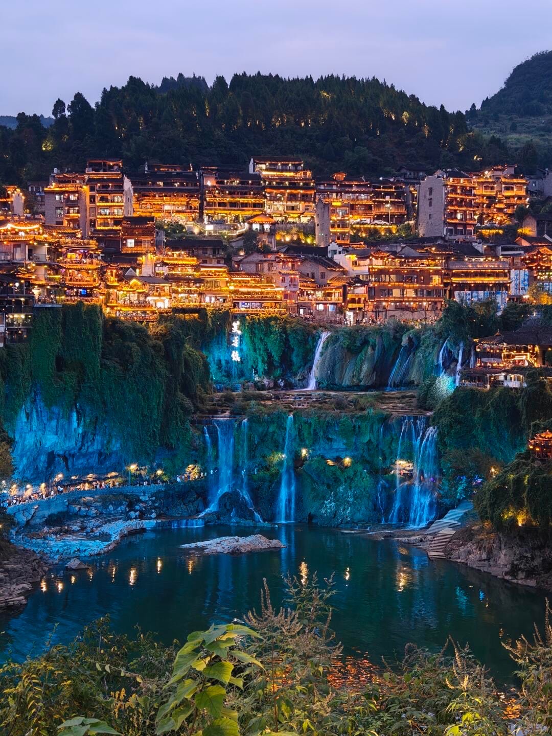 Cliffside old town glowing with golden lights above a floodlit waterfall tumbling into a river pool at dusk.